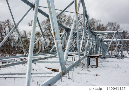 Galvanized steel power line tower sections lying on a snow-covered field, awaiting assembly. Bare trees and a cloudy winter sky complete the scene 140041125