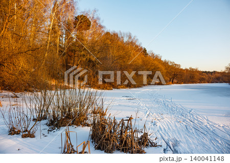 Golden light bathes a serene frozen river, its snowy expanse marked by ski tracks. Bare trees line the bank, adding a warm glow to the tranquil winter landscape Golden light bathes a serene frozen river, its snowy expanse marked by ski tracks. Bare trees line the bank, adding a warm glow to the tranquil winter landscape 140041148