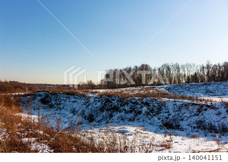 Winter landscape featuring an old, snow-dusted quarry with dry grass and a distant line of bare trees under a clear blue sky. A tranquil, frosty scene 140041151