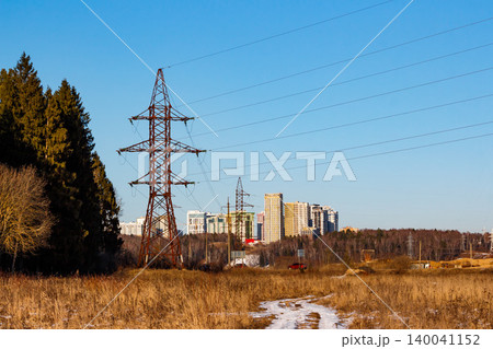 Gnarly pylon stands tall in a dry field under clear blue skies, backed by a sprawling town and distant woods. Rural landscape meets power lines 140041152