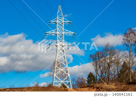 Towering electricity pylon dominates a clear blue sky, laced with high-voltage cables. Leafless trees dot the sun-drenched, earthy terrain below, showcasing essential infrastructure 140041161