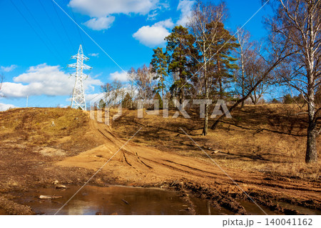 A prominent power line pylon towers over a rugged dirt track snaking uphill, framed by leafless trees and a muddy puddle under a bright blue sky 140041162