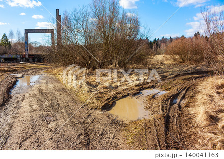 Gnarly dirt track with puddles cuts through a barren field under a bright sky, hinting at challenging travel near forgotten industrial structures 140041163