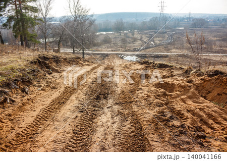 Rough sandy track, deeply etched with tire marks, slopes downwards. A muted, wintry panorama unfolds, featuring sparse trees, open fields, and a distant power pylon 140041166