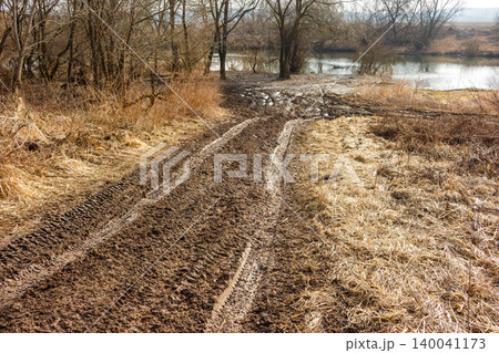 Muddy off-road trail, deeply rutted, meanders through withered grass towards a tranquil river. Bare trees line the water's edge, suggesting early spring scenery Muddy off-road trail, deeply rutted, meanders through withered grass towards a tranquil river. Bare trees line the water's edge, suggesting early spring scenery 140041173