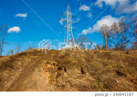 A robust power line pylon stands tall on a dry hillside under a bright blue sky with sparse clouds. Industrial landscape meets nature's raw beauty A robust power line pylon stands tall on a dry hillside under a bright blue sky with sparse clouds. Industrial landscape meets nature's raw beauty 140041187