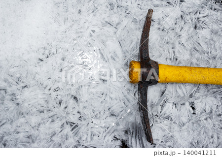 Rustic pickaxe with bright yellow handle rests on a frosty expanse of intricate ice crystals and fresh snow. A scene of winter resilience or cold endeavor 140041211