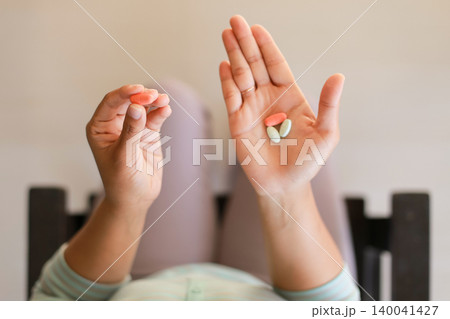Hands hold two pills, one pink, one green. Person rests in a cozy, softly lit room. Calm demeanor suggests routine or health care. Perfect for wellness, medicine, or health Hands hold two pills, one pink, one green. Person rests in a cozy, softly lit room. Calm demeanor suggests routine or health care. Perfect for wellness, medicine, or health 140041427