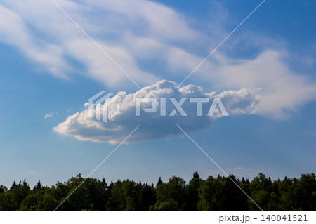Single fluffy cumulus cloud floats in a vibrant azure sky above a lush green forest line, creating a peaceful atmospheric scenery on a bright sunny summer day 140041521