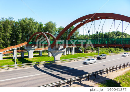 Modern arched pedestrian bridge over a multi-lane highway with moving cars, surrounded by lush green trees on a sunny day providing urban transportation infrastructure Modern arched pedestrian bridge over a multi-lane highway with moving cars, surrounded by lush green trees on a sunny day providing urban transportation infrastructure 140041875