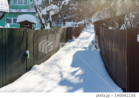 Sun-kissed snowy alley between corrugated fences, a serene winter wonderland leading to distant cozy village homes and frosted trees Sun-kissed snowy alley between corrugated fences, a serene winter wonderland leading to distant cozy village homes and frosted trees 140042188
