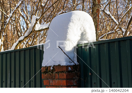 Thick snowdrift caps a brick fence post beside a dark green corrugated barrier, winter forest backdrop Thick snowdrift caps a brick fence post beside a dark green corrugated barrier, winter forest backdrop 140042204