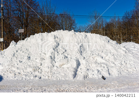 Massive snowdrift, cleared by machinery, dominates the urban street with bare trees and clear blue sky in background 140042211