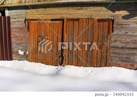 Weathered orange wooden garage doors stand buried in deep snow drifts against a rustic plank wall Weathered orange wooden garage doors stand buried in deep snow drifts against a rustic plank wall 140042333