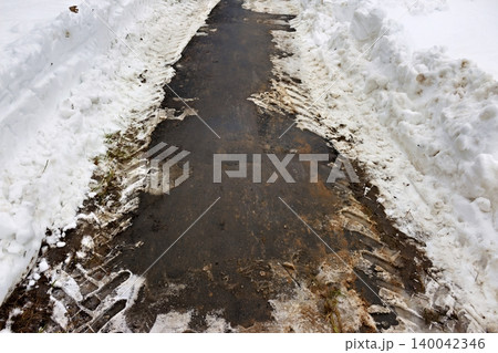 Dirty slushy snow piled along a cleared pathway after winter plowing, showing tire tracks and wet asphalt 140042346