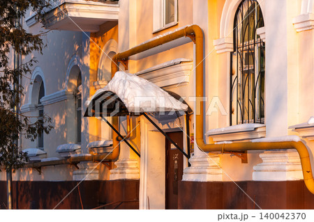 Golden hour sunlight bathes a classic building facade, highlighting a snow-capped canopy and industrial pipes. Winter charm meets urban warmth Golden hour sunlight bathes a classic building facade, highlighting a snow-capped canopy and industrial pipes. Winter charm meets urban warmth 140042370