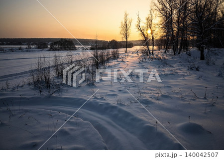 Golden hour casts a warm glow over a serene winter wonderland. Freshly fallen snow blankets the peaceful landscape, with bare trees silhouetted against the vibrant sunset sky Golden hour casts a warm glow over a serene winter wonderland. Freshly fallen snow blankets the peaceful landscape, with bare trees silhouetted against the vibrant sunset sky 140042507