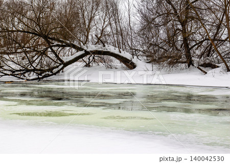 Fragile, melting river ice shimmers with hints of green, a clear sign of late winter's slow thaw. Snow blankets banks, skeletal trees line the background Fragile, melting river ice shimmers with hints of green, a clear sign of late winter's slow thaw. Snow blankets banks, skeletal trees line the background 140042530
