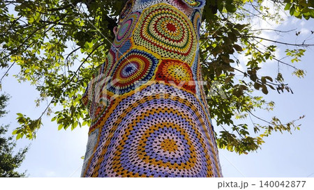 A tree trunk wrapped in colorful knitted cover stands on city street. Handmade yarn bombing installation transforms natural surface into vibrant piece of urban art, blending creativity and texture A tree trunk wrapped in colorful knitted cover stands on city street. Handmade yarn bombing installation transforms natural surface into vibrant piece of urban art, blending creativity and texture 140042877