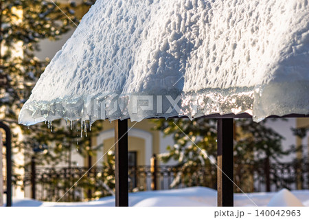Melting snow and ice overhang on a roof edge with sunlit icicles dripping in the late winter or early spring thaw Melting snow and ice overhang on a roof edge with sunlit icicles dripping in the late winter or early spring thaw 140042963