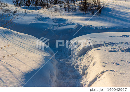 Deeply carved snow path winding through high drifts under bright winter sunlight, casting long blue shadows across the pristine white mounds Deeply carved snow path winding through high drifts under bright winter sunlight, casting long blue shadows across the pristine white mounds 140042967