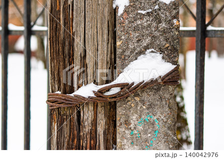 Old wooden post tightly hugged by a concrete pillar and rusty metal cable, topped with fresh winter snow. Grungy outdoor close-up Old wooden post tightly hugged by a concrete pillar and rusty metal cable, topped with fresh winter snow. Grungy outdoor close-up 140042976