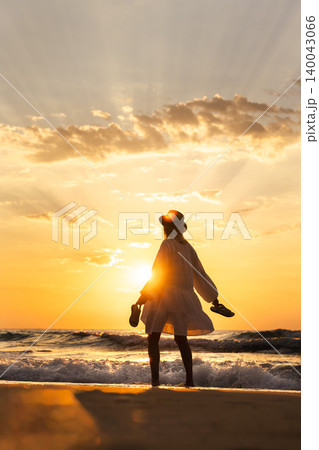 Middle aged woman in hat standing by sea waves at sunrise holding shoes, freedom travel lifestyle concept 140043066