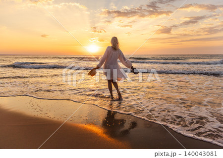 Middle aged woman standing in sea waves at sunrise holding hat and shoes, freedom travel lifestyle concept Middle aged woman standing in sea waves at sunrise holding hat and shoes, freedom travel lifestyle concept 140043081