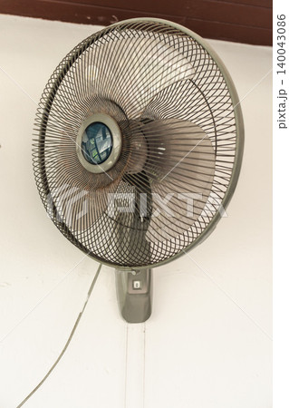 Wall mounted electric fan with metal grille on white interior wall, household cooling device for air circulation during hot weather Wall mounted electric fan with metal grille on white interior wall, household cooling device for air circulation during hot weather 140043086