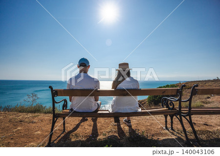 Couple sitting on bench overlooking Black Sea coast under bright summer sun in Bulgaria Couple sitting on bench overlooking Black Sea coast under bright summer sun in Bulgaria 140043106