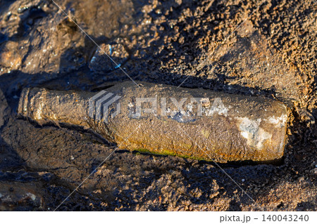 Weathered glass bottle coated in thick mud resting on a dark sandy beach shore, reminiscent of a mysterious message found washed up by the ocean tide during a sunny day Weathered glass bottle coated in thick mud resting on a dark sandy beach shore, reminiscent of a mysterious message found washed up by the ocean tide during a sunny day 140043240