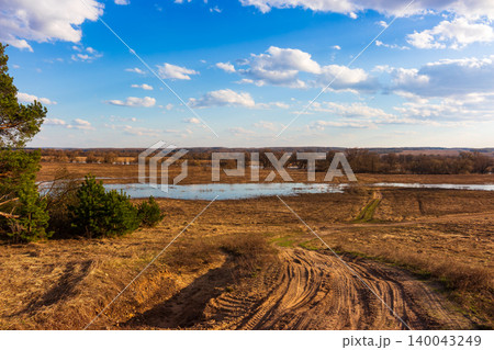 Dirt road winds through a dry meadow toward a spring flooded riverbank under a bright blue sky with fluffy white clouds on a serene sunny day in the countryside Dirt road winds through a dry meadow toward a spring flooded riverbank under a bright blue sky with fluffy white clouds on a serene sunny day in the countryside 140043249