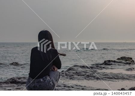海辺の岩場で静かに海を見つめる女性の後ろ姿 海辺の岩場で静かに海を見つめる女性の後ろ姿 140043457