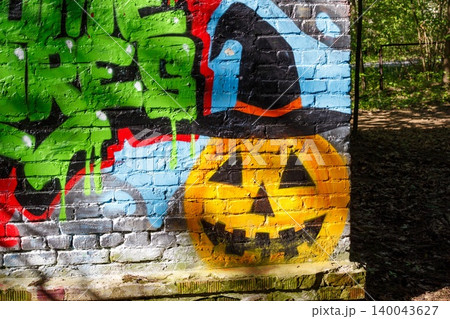 Vibrant street art features a spooky jack-o'-lantern face with a witch hat painted on a weathered brick wall next to wild green graffiti: Vibrant street art features a spooky jack-o'-lantern face with a witch hat painted on a weathered brick wall next to wild green graffiti: 140043627