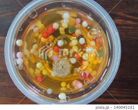 Fungus on the surface of children's yellow fruit jelly with colorful balls in a plastic cup, poor-quality poisonous snack, spoilage of sweets due to improper storage, mold on a gelatin product Fungus on the surface of children's yellow fruit jelly with colorful balls in a plastic cup, poor-quality poisonous snack, spoilage of sweets due to improper storage, mold on a gelatin product 140045281