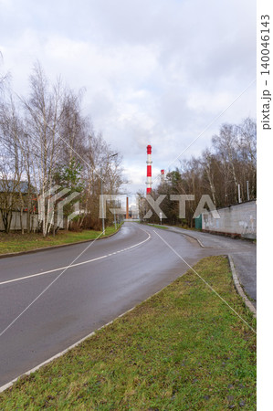 Quiet road alongside factory smoke stacks. Bare trees and reflective wet road under gloomy sky Quiet road alongside factory smoke stacks. Bare trees and reflective wet road under gloomy sky 140046143