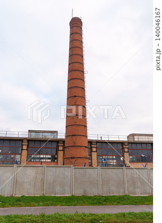 Ruined factory with tall weathered brick chimney standing silently against cloudy grey sky 140046167