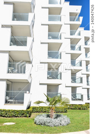 Contemporary apartment building features multiple balconies with glass railings, surrounded by a well-maintained garden with palm trees and shrubs, under a clear blue sky Contemporary apartment building features multiple balconies with glass railings, surrounded by a well-maintained garden with palm trees and shrubs, under a clear blue sky 140047426