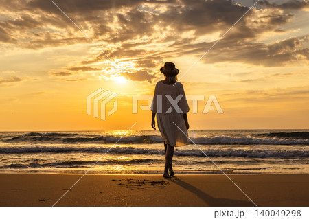 Middle aged woman walking on beach toward sea at sunrise, peaceful freedom travel lifestyle concept Middle aged woman walking on beach toward sea at sunrise, peaceful freedom travel lifestyle concept 140049298