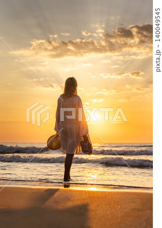 Middle aged woman standing on beach at sunrise holding hat and shoes, freedom travel lifestyle by sea Middle aged woman standing on beach at sunrise holding hat and shoes, freedom travel lifestyle by sea 140049545