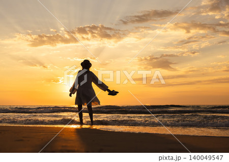 Middle aged woman standing in sea waves at sunrise with shoes in hand, freedom travel lifestyle concept Middle aged woman standing in sea waves at sunrise with shoes in hand, freedom travel lifestyle concept 140049547