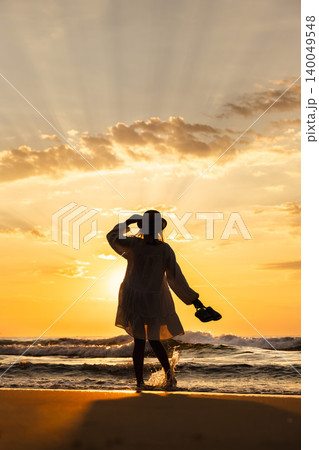 Middle aged woman in hat standing by sea at sunrise holding shoes, freedom travel lifestyle concept Middle aged woman in hat standing by sea at sunrise holding shoes, freedom travel lifestyle concept 140049548