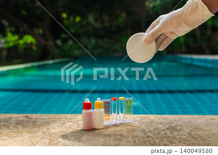 Gloved hand holding round chlorine tablet above swimming pool edge with water test kit and reagent bottles, Gloved hand holding round chlorine tablet above swimming pool edge with water test kit and reagent bottles, 140049580