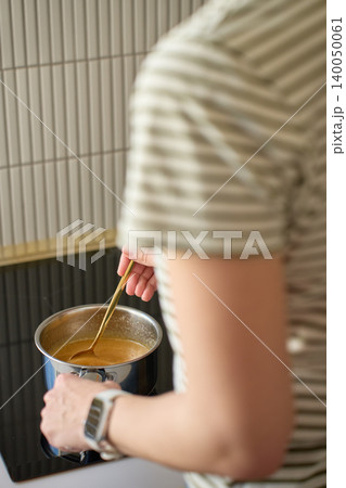 Woman stirs mixture in metal saucepan on kitchen stovetop Woman stirs mixture in metal saucepan on kitchen stovetop 140050061