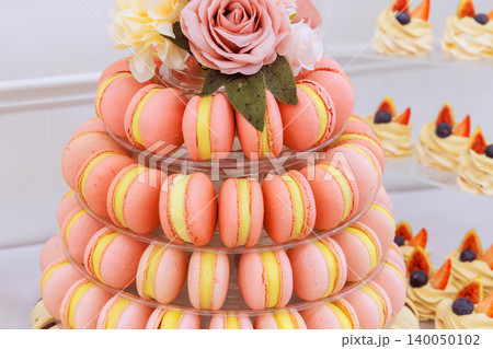 Bright tower displays various pink, yellow macarons with flower on top at dessert table during celebration Bright tower displays various pink, yellow macarons with flower on top at dessert table during celebration 140050102