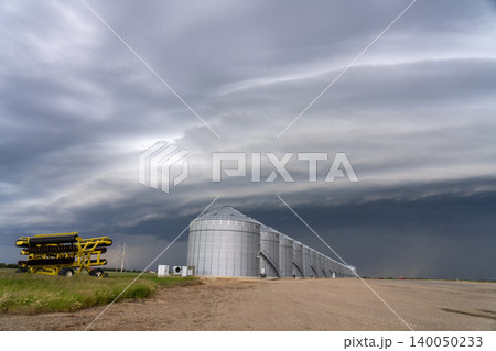Agricultural grain silos stand under a dramatic, stormy sky with a farm implement nearby 140050233