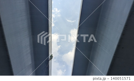 Low angle perspective captures massive twin concrete overpass structures, framing a bright blue sky filled with soft white clouds. The heavy minimalist urban architecture creates strong parallel 140051571