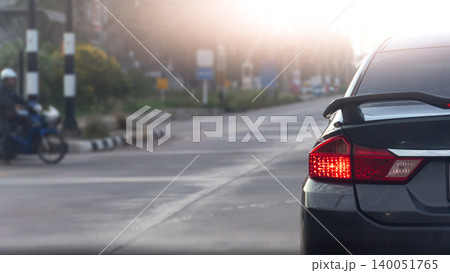 Rear corner close up of a dark car showing its illuminated red tail light. Car is stopped on a wide city road with soft daylight. Blurred of motorcycles waiting to cross the intersection. 140051765