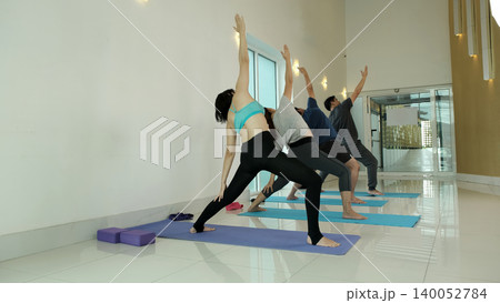 Group of people practicing reverse warrior yoga pose in bright modern studio, stretching side body and opening chest to improve flexibility, balance and strength in guided fitness and wellness class 140052784