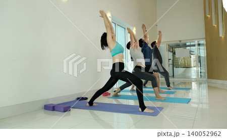 Group of people practicing warrior yoga pose in bright modern studio, stretching legs and opening chest to improve balance, strength and flexibility during guided fitness and wellness class Group of people practicing warrior yoga pose in bright modern studio, stretching legs and opening chest to improve balance, strength and flexibility during guided fitness and wellness class 140052962